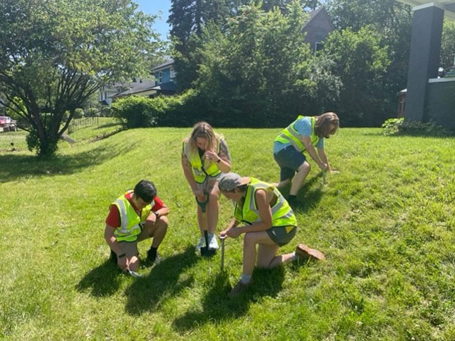 Four students in safety vests use small shovels to dig up soil samples in a grassy field.